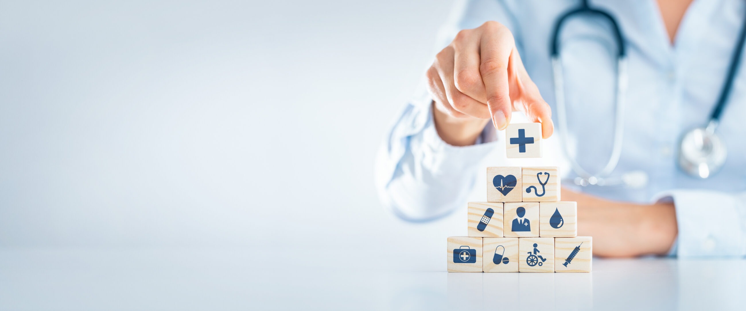 A healthcare professional with a stethoscope places a wooden block with a medical cross on top of a pyramid of blocks featuring healthcare icons symbolizing the importance of building a layered asset protection strategy to safeguard a medical practice.
