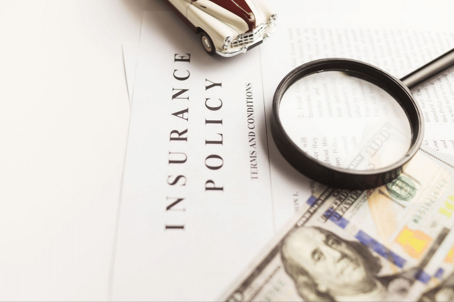 Insurance policy document with a magnifying glass and a $100 bill on a white desk.