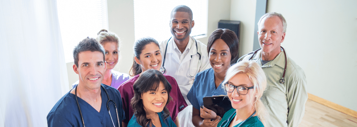 A diverse group of smiling medical professionals including doctors and nurses in scrubs and white coats gathered together, representing the hardworking healthcare teams who benefit from asset protection strategies designed for medical professionals.