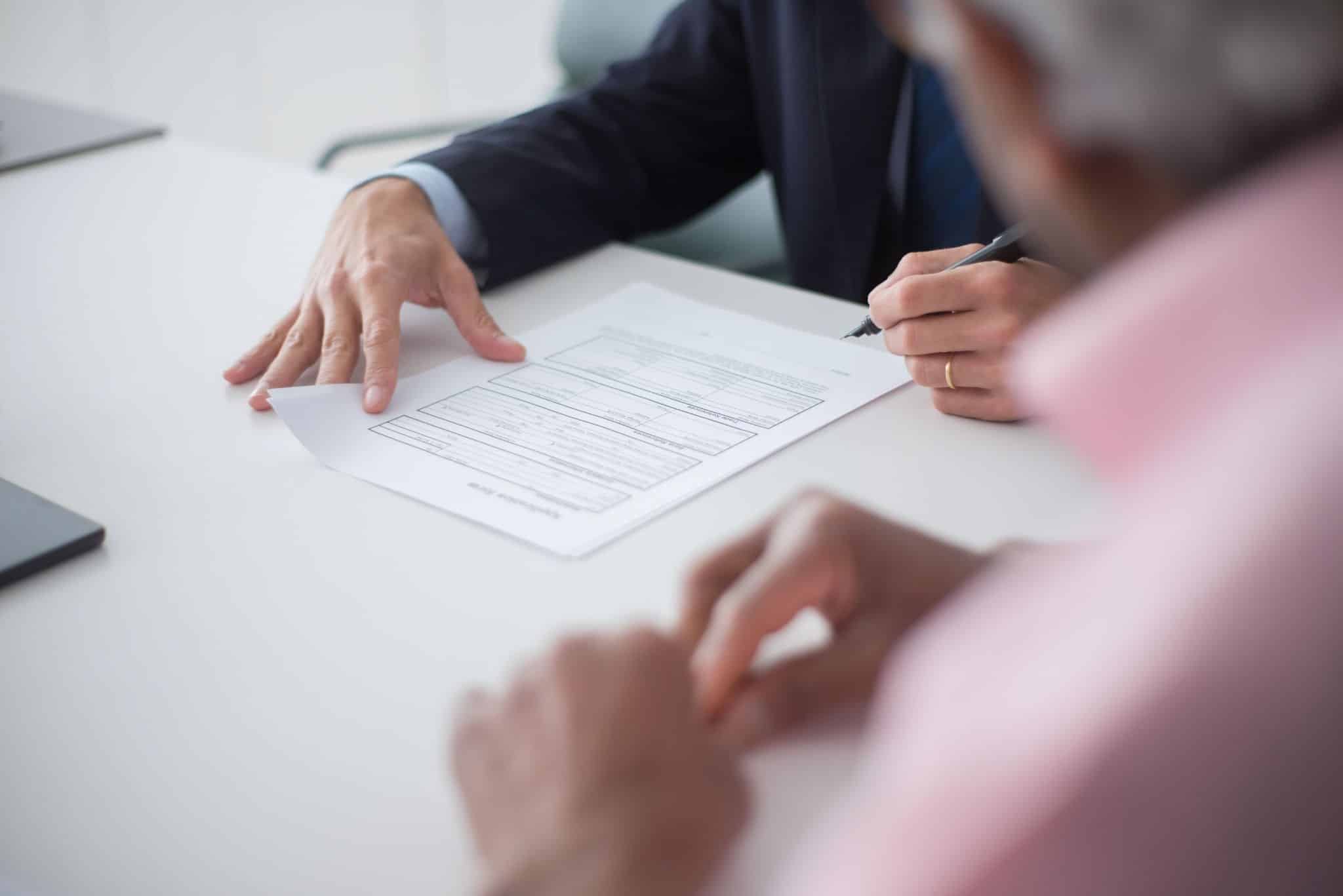 A person in a dark suit sits at a white table, holding a pen over a printed application form. They are viewed over the shoulder of an out-of-focus person wearing a pink shirt. 
