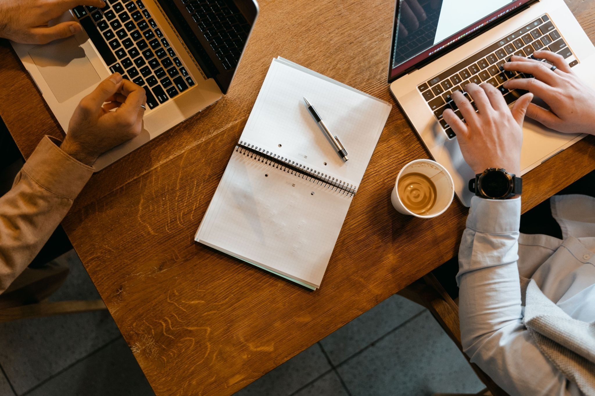 Overhead view of two people working on laptops, with coffee and a notebook on the desk.