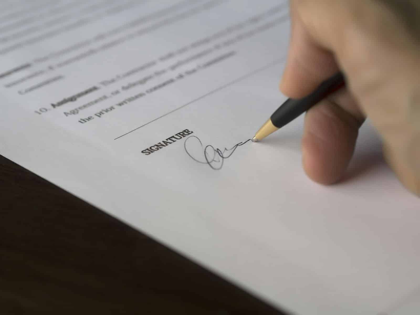 A close-up shot of a hand using a black and gold pen to sign a legal document on a line marked "SIGNATURE."