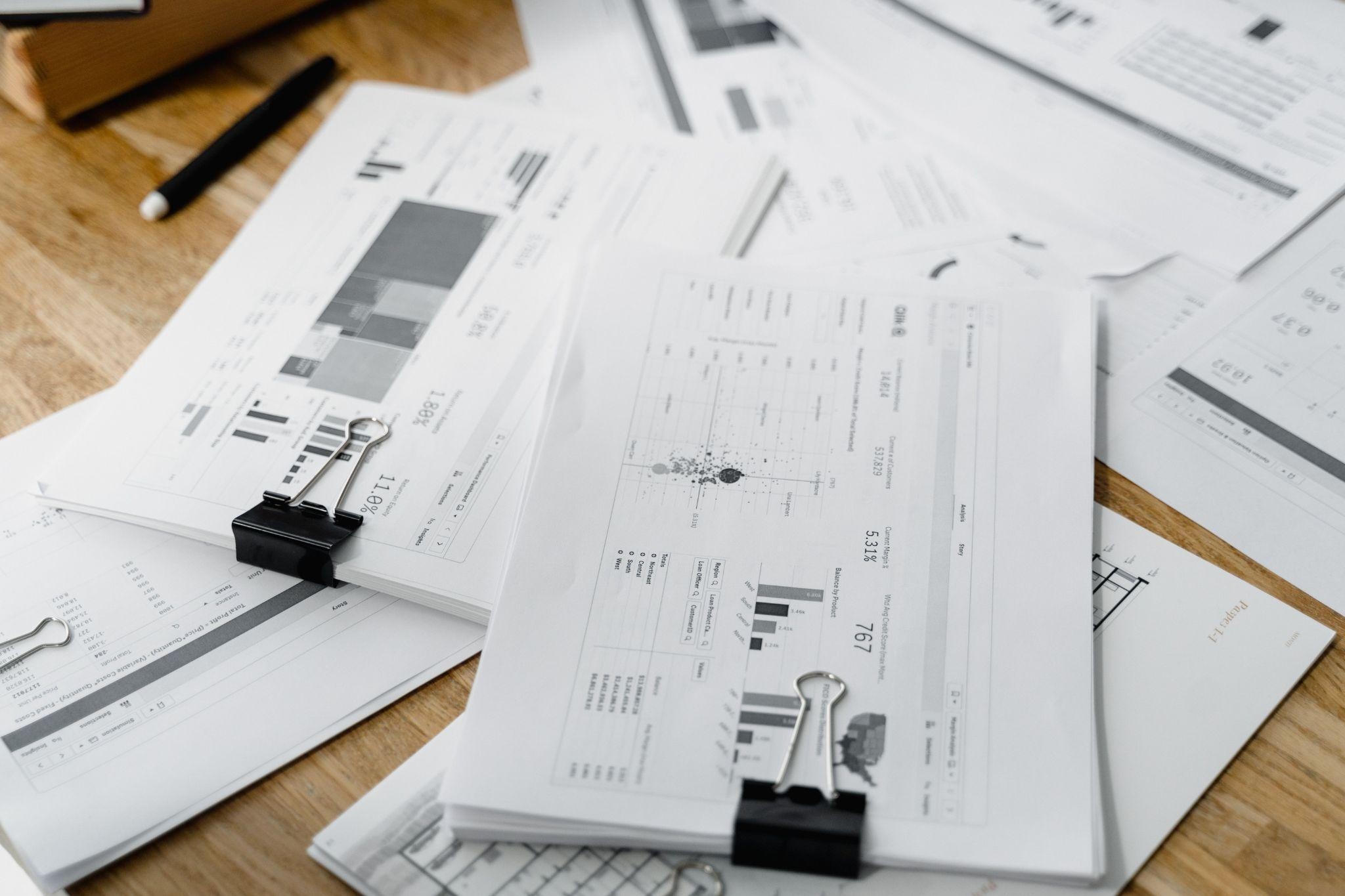 Printed charts and financial reports held with binder clips on a wooden desk
