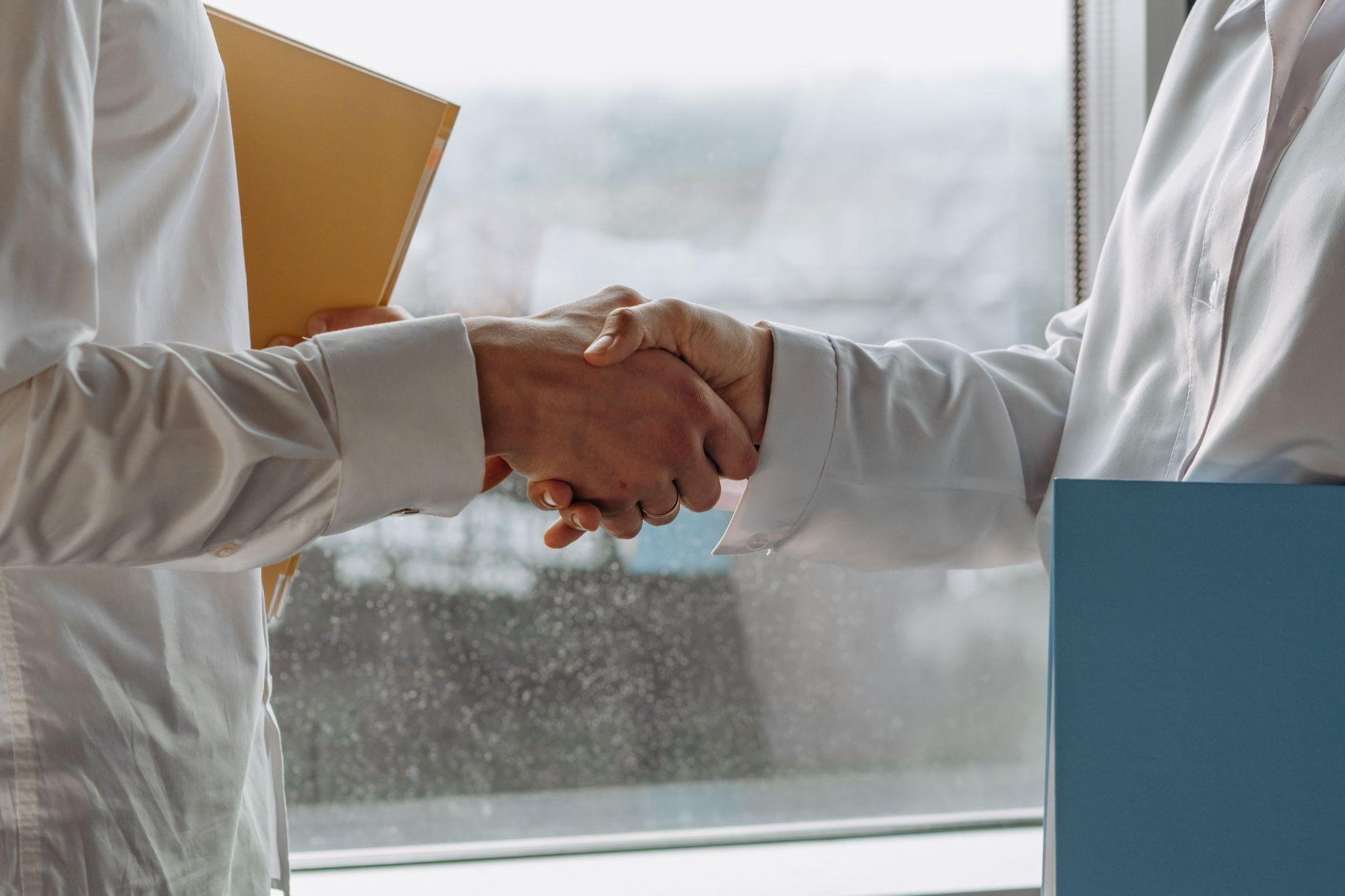 Two professionals in white shirts shaking hands while holding folders
