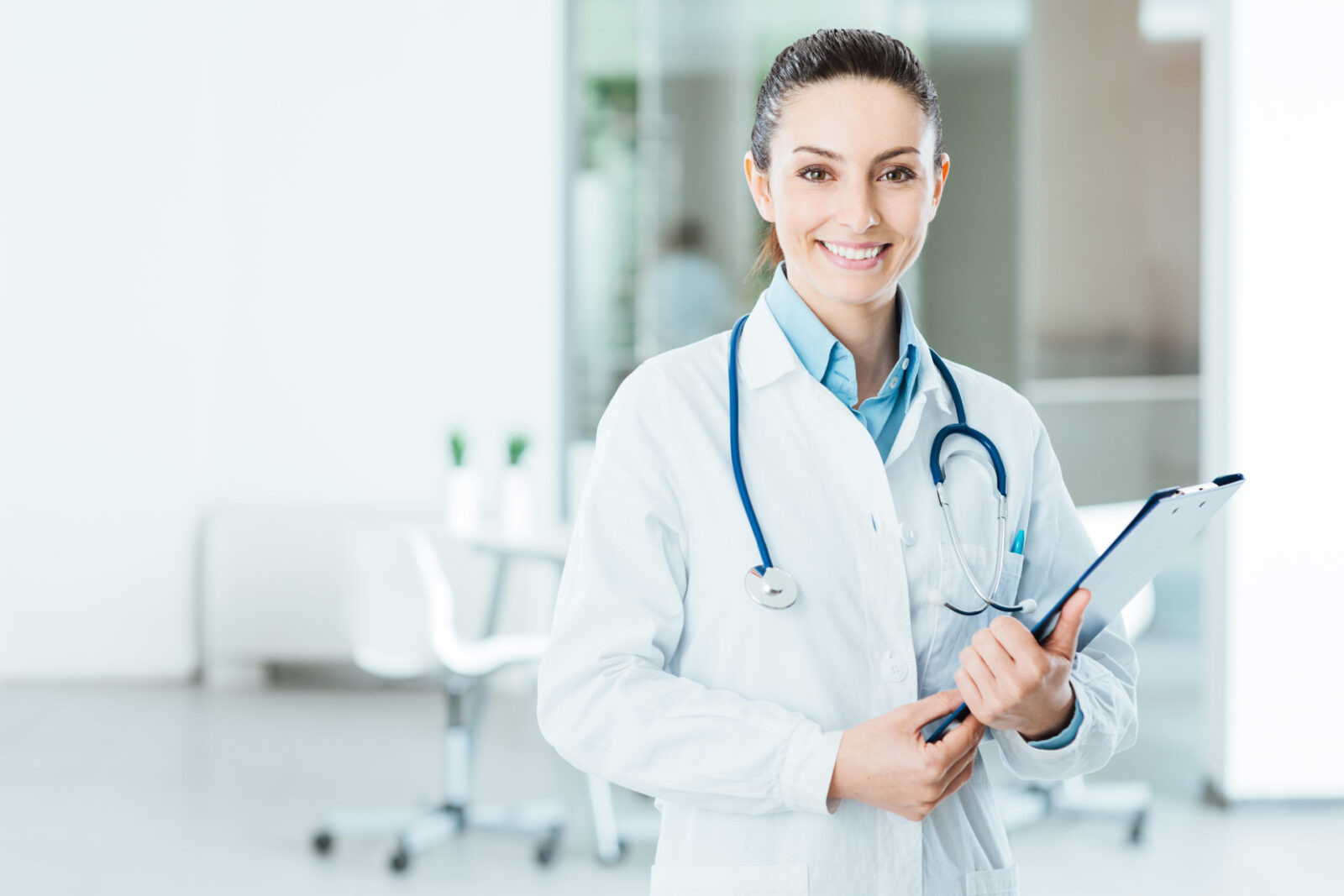 A confident female physician in a white coat and stethoscope holding a clipboard in a medical office, representing the high-earning medical professionals who need asset protection strategies beyond professional liability insurance.
